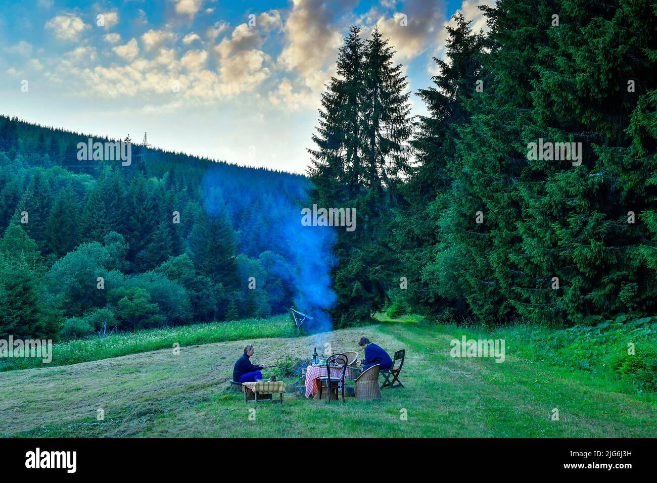 two persons sitting by campfire in summer evening in the mountain country Stock Photo