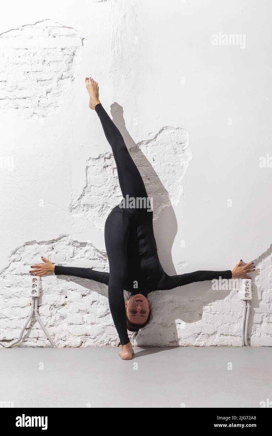 A woman in a black sports overalls performs exercises for stretching the muscles of the legs, doing a longitudinal split, leaning her back against the Stock Photo