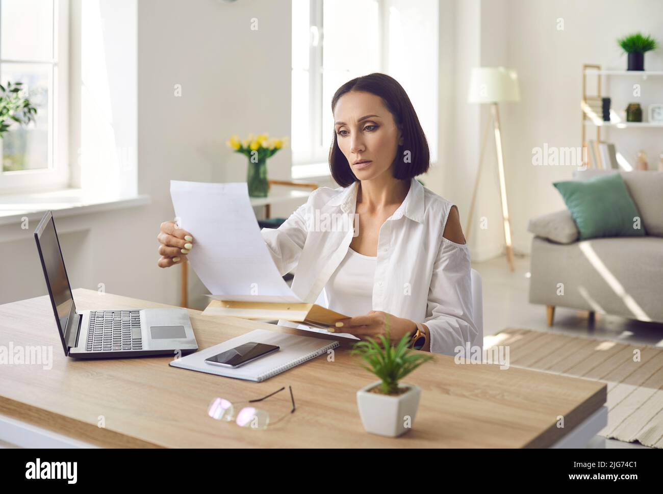 Serious adult woman sitting at her desk and reading letter, bill or notice of debt Stock Photo