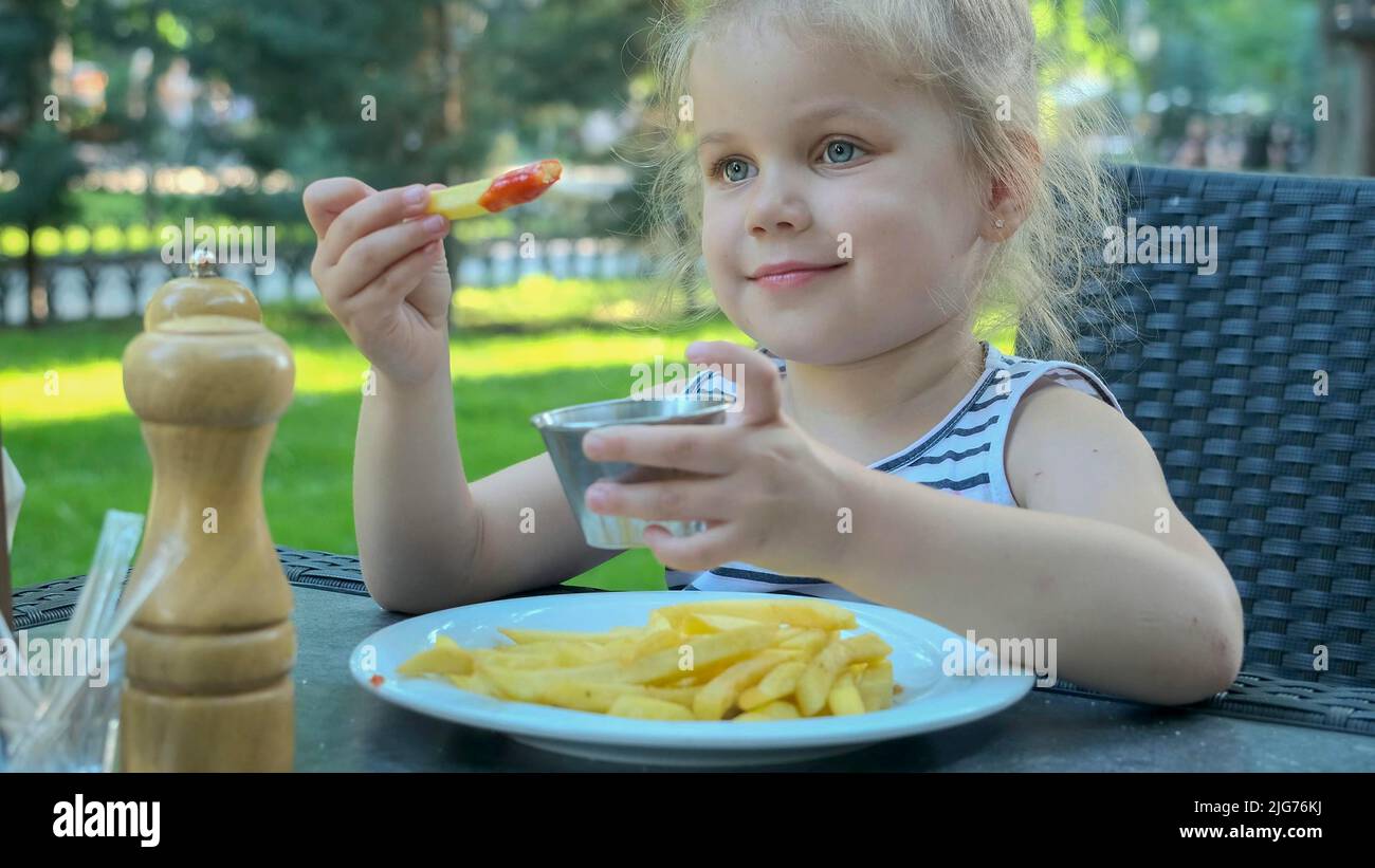 Little girl eat french fries. Close-up of blonde girl takes potato chips with her hands and tries them sitting in street cafe on the park. Odessa Stock Photo