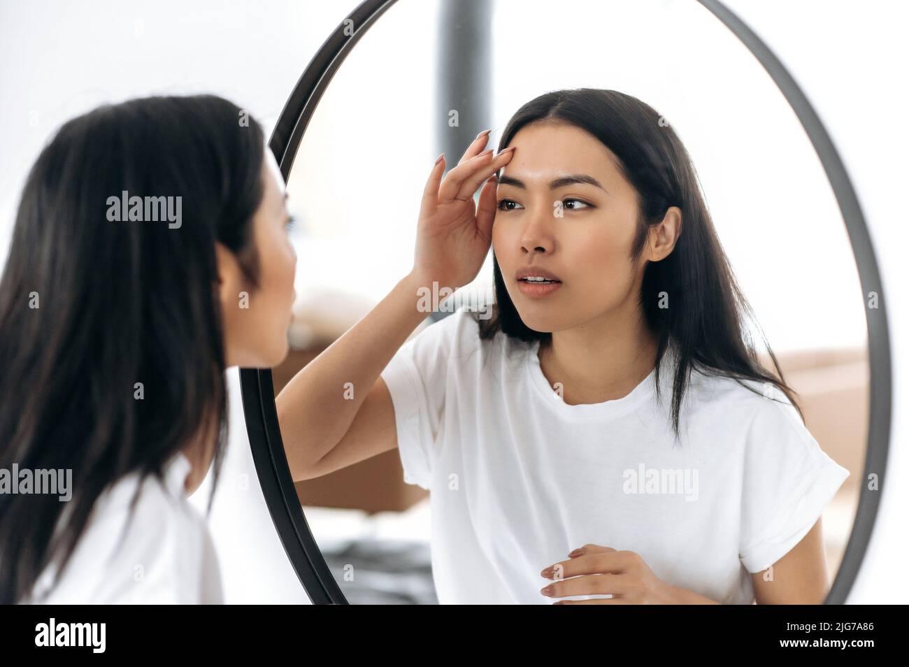 Satisfied lovely chinese girl with long brunette hair in white t-shirt, stands in front of a mirror, examines her face, happy at the healthy state of her facial skin, smile, getting ready to do makeup Stock Photo