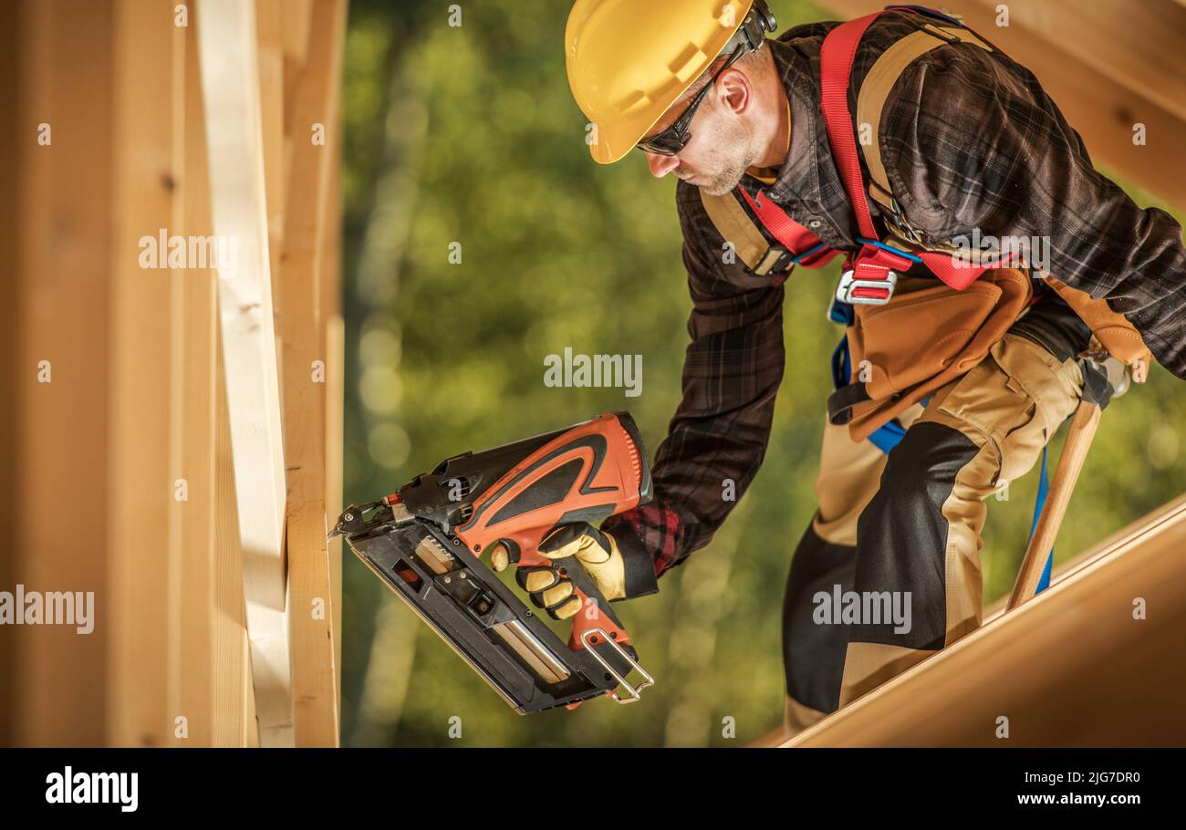 Caucasian Construction Worker with a Nail Gun Assembling Wooden Skeleton Frame Beams in a Newly Developed Building. Construction Industry Theme. Stock Photo