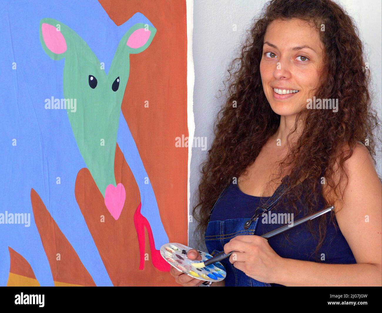 Smiling beautiful young female Mexican painter (visual artist) with long curly hair holds brush and palette in front of her painting. Stock Photo