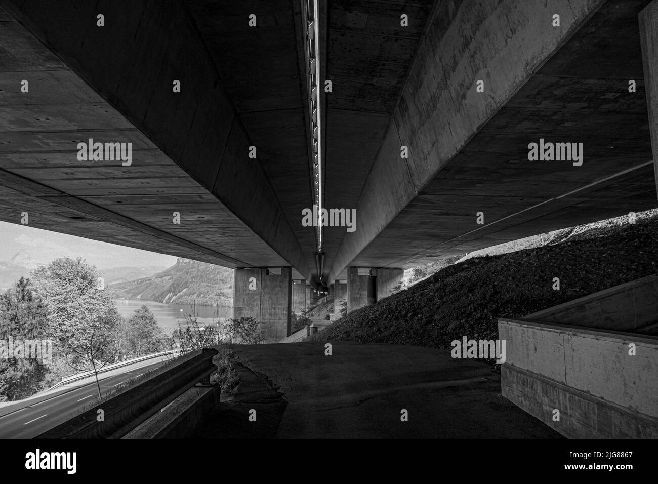 A grayscale of a road under the massive concrete bridge by a river Stock Photo