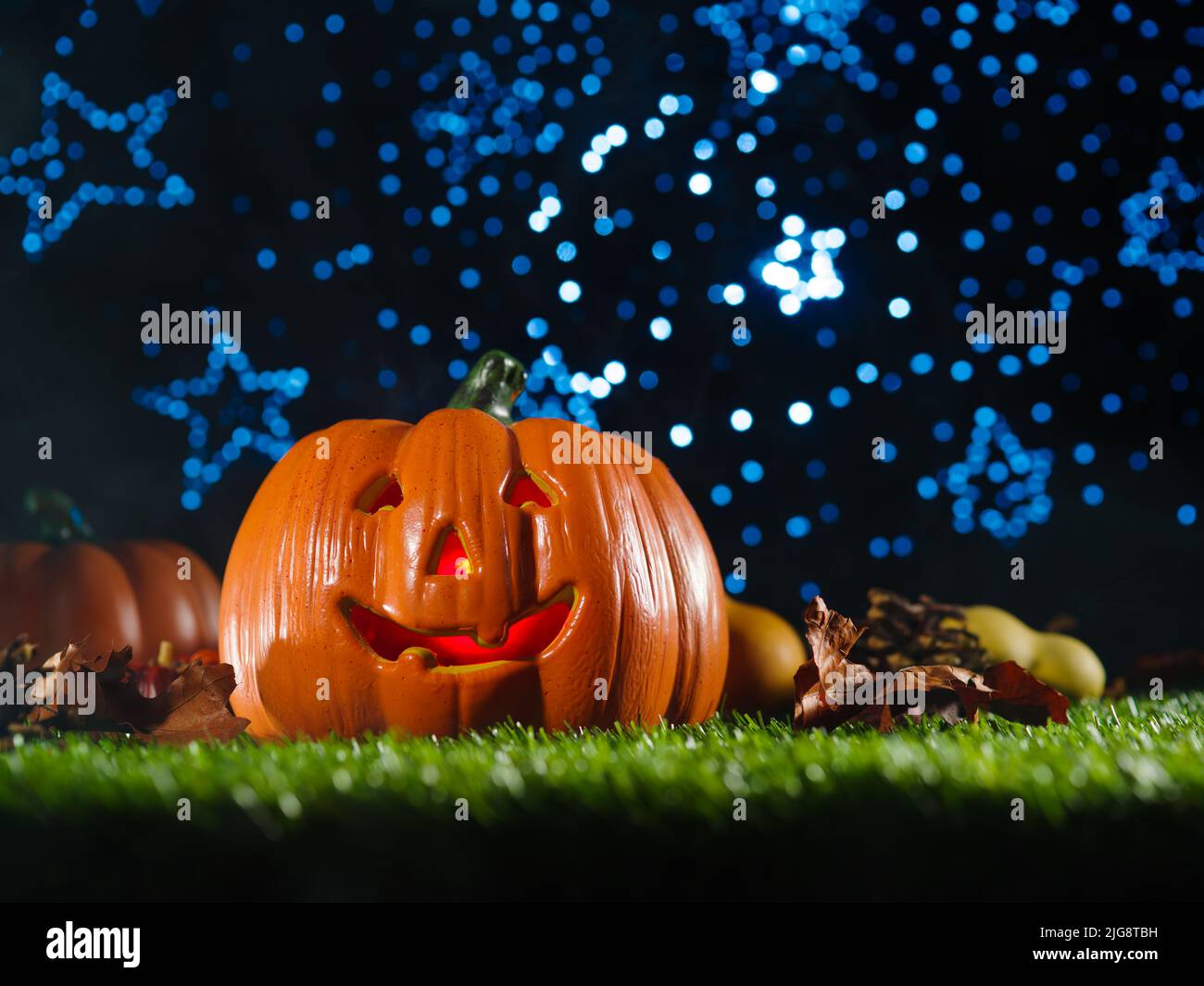 Mystical Halloween composition. Smiling halloween pumpkin with illuminated carved face, autumn fruits, cones on a green lawn against a blue shimmering Stock Photo