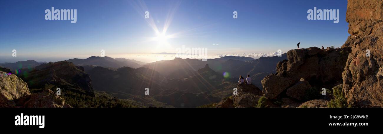 Spain, Canary Islands, Gran Canaria, Massif Central, Roque Nublo, panorama shot, sunset, sun as star, blue sky, view over Massif Central to the west, Teide, Tenerife, couple sitting from behind, two people next to the Roque Stock Photo