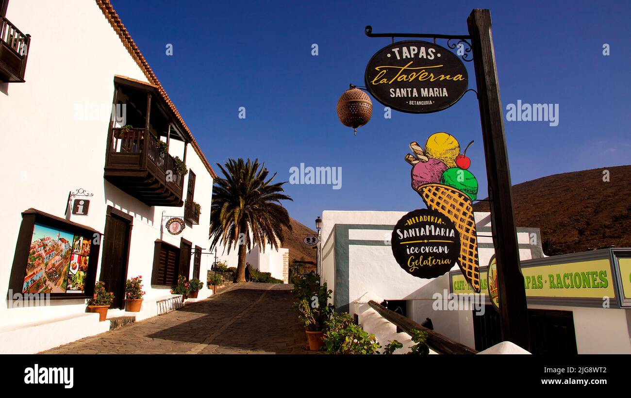Spain, Canary Islands, Fuerteventura, Betancuria, old capital, old town, alley, wide angle shot, left white row of houses, right sign of an ice cream parlor, blue cloudless sky Stock Photo