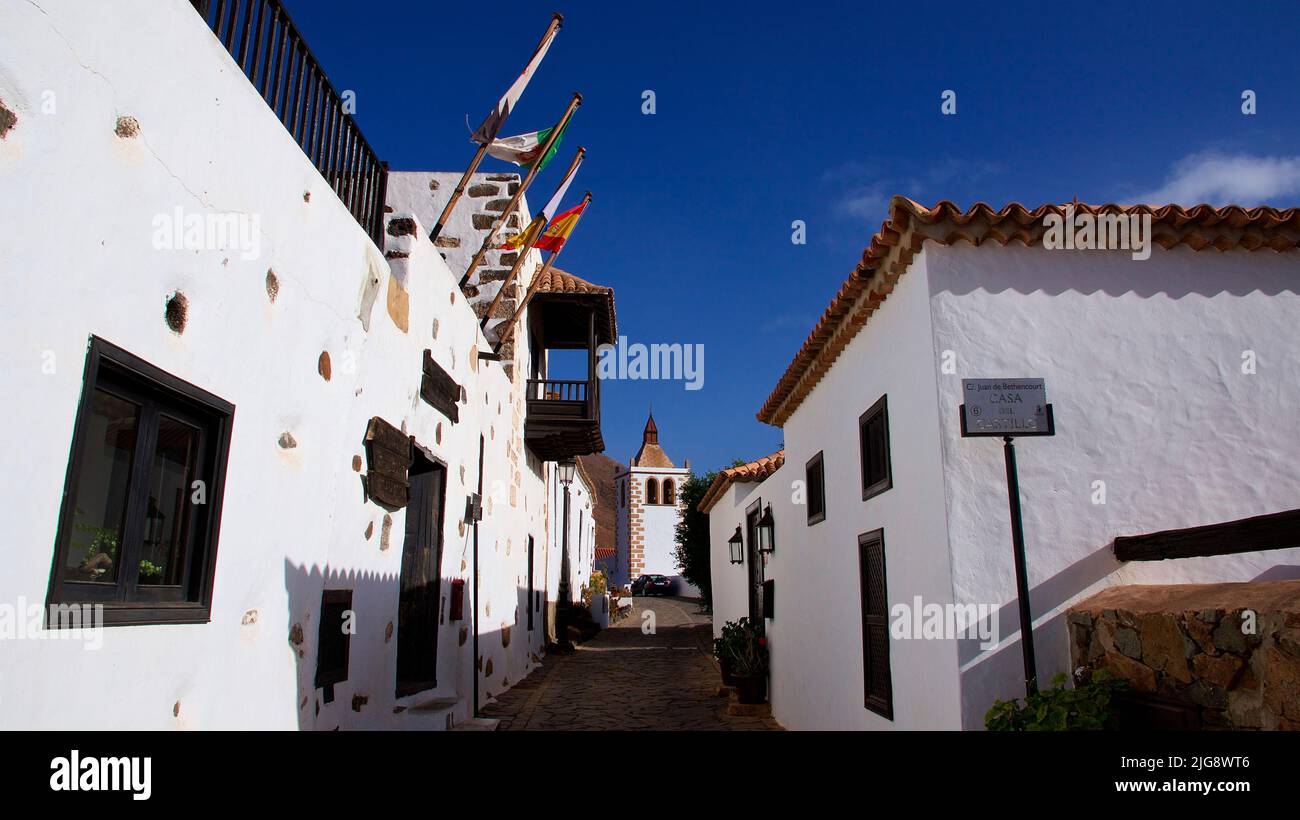 Spain, Canary Islands, Fuerteventura, Betancuria, old capital, old town, alley, church at the end of the alley, flags, white houses, wooden balcony, blue sky, scattered white clouds Stock Photo