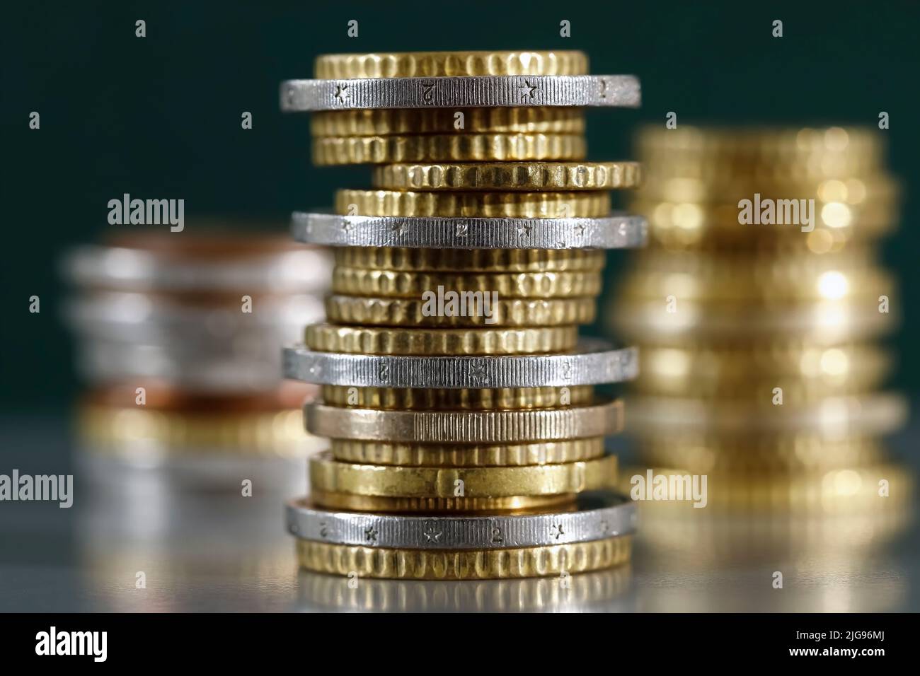 The stacked euro coins have been placed on a grey, flat surface and on a dark green background. There are three piles of coins. Stock Photo