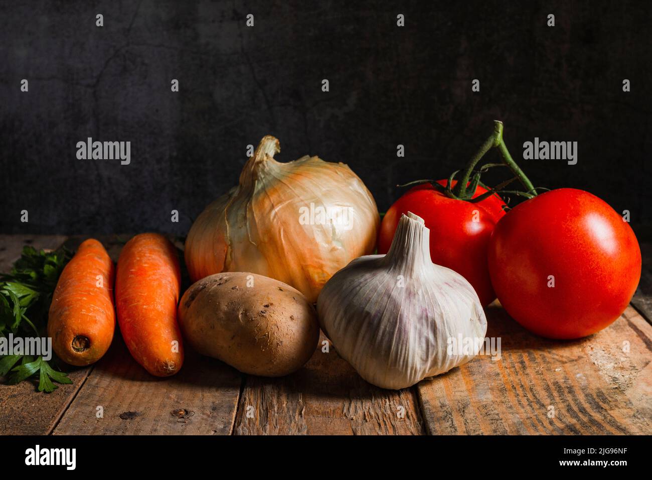 Still life on a rustic table of wooden planks on which there are carrots, tomatoes, garlic, onion, potato and parsley. In the background there is a wa Stock Photo