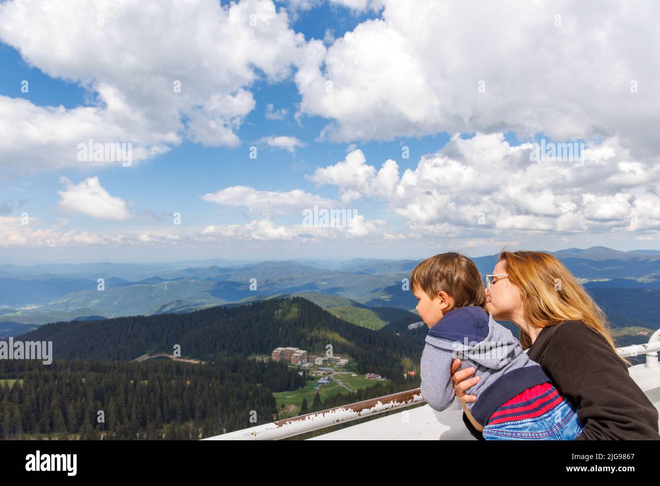 Mother holds her baby son tightly and shows him the landscapes of mountain forest valley of the Rhodope Mountains and the blue cloudy sky, while stand Stock Photo