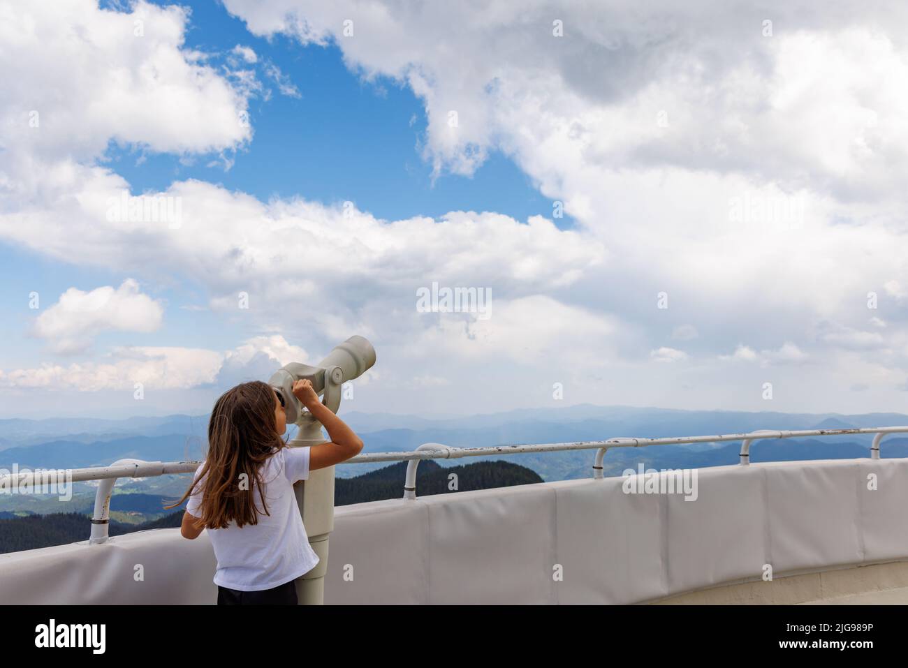 Curious little girl looking at landscapes of mountain forest valley of Rhodope Mountains and bright blue cloudy sky through modern metal telescope, on Stock Photo