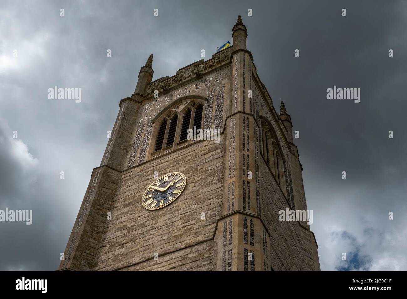 The tower of All Saints Church Laxfield against an angry dark sky with the clock face glinting in the sunlight Stock Photo