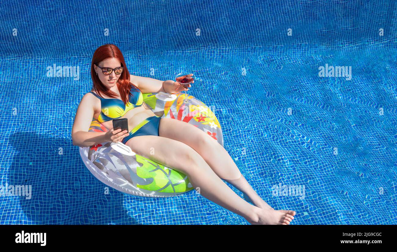 young red-haired girl, looking at her smartphone floating on a ring float inside the pool, while holding a soft drink. young girl on holiday relaxing Stock Photo