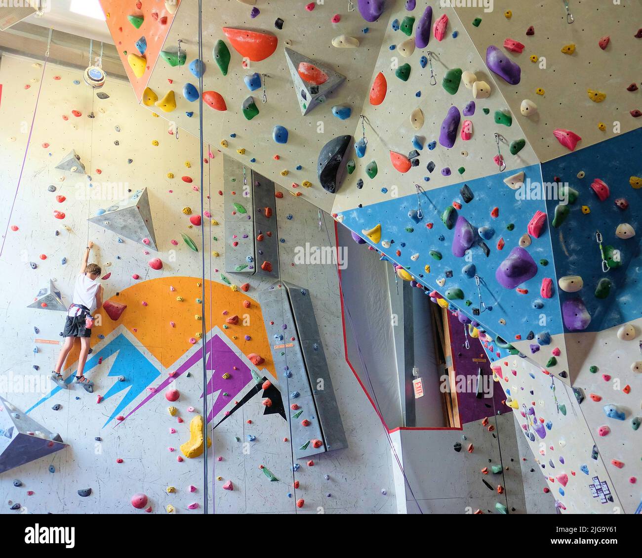 Young teenager climbing on an Indoor rock climbing wall seen from below; ropes and climbing holds for support on the ascent. Stock Photo