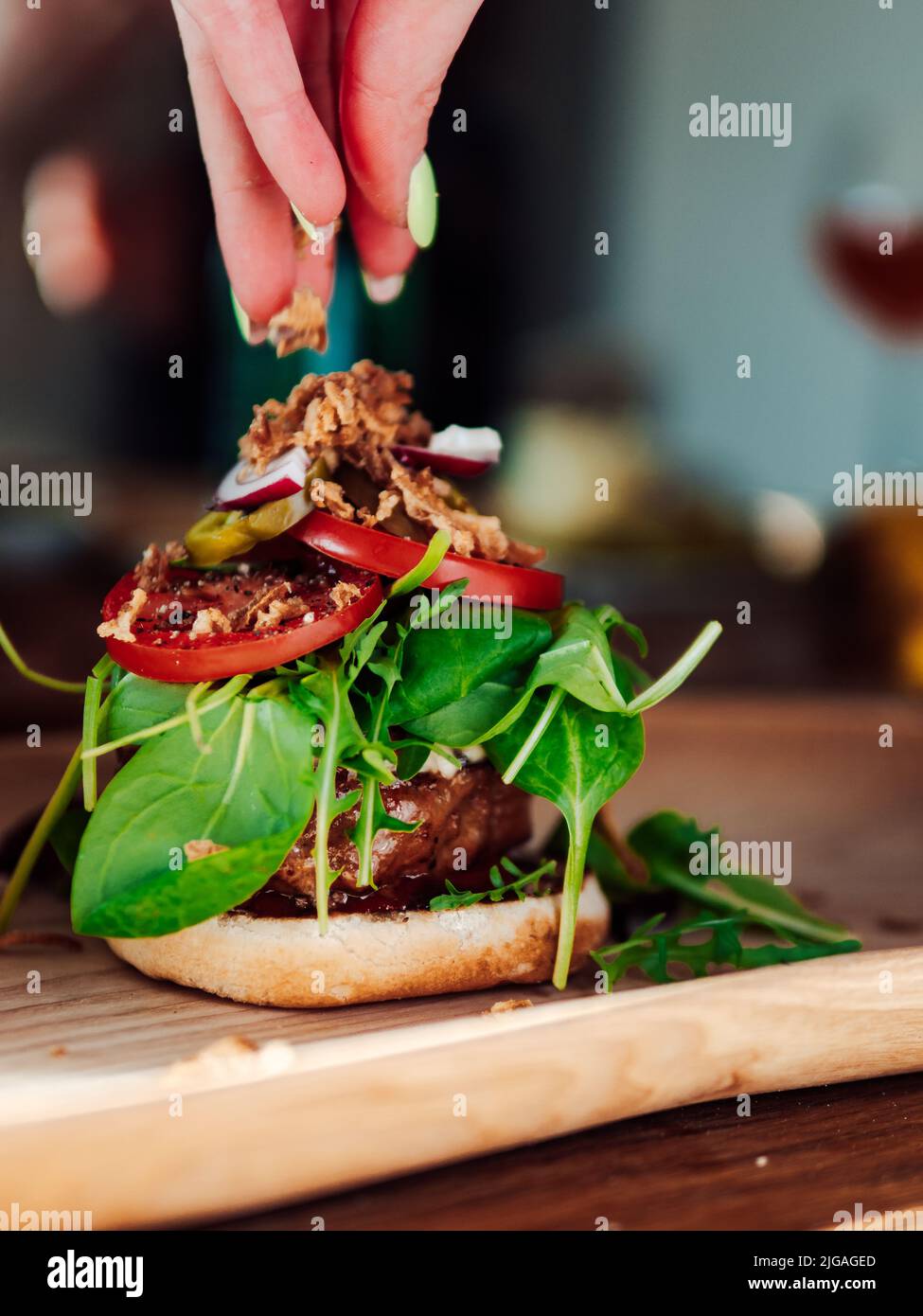 A female hand dressing a yummy burger with meat, salad and other ingredients on a wooden surface Stock Photo