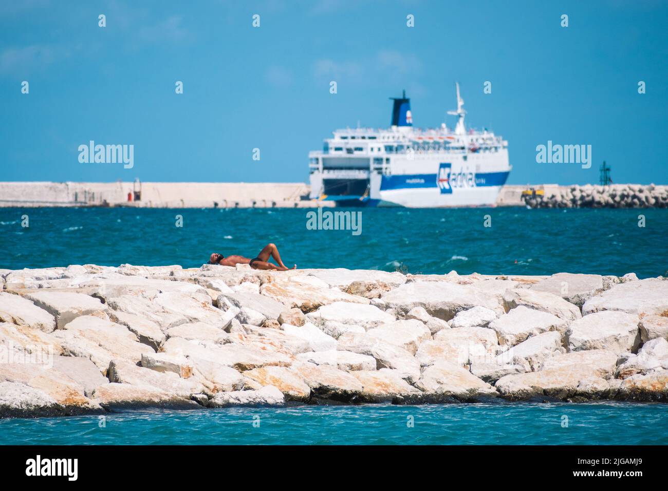 Tanned bather sunbathing on the breakwater with pier, cruise ship, blue sky, blue sea on background Stock Photo