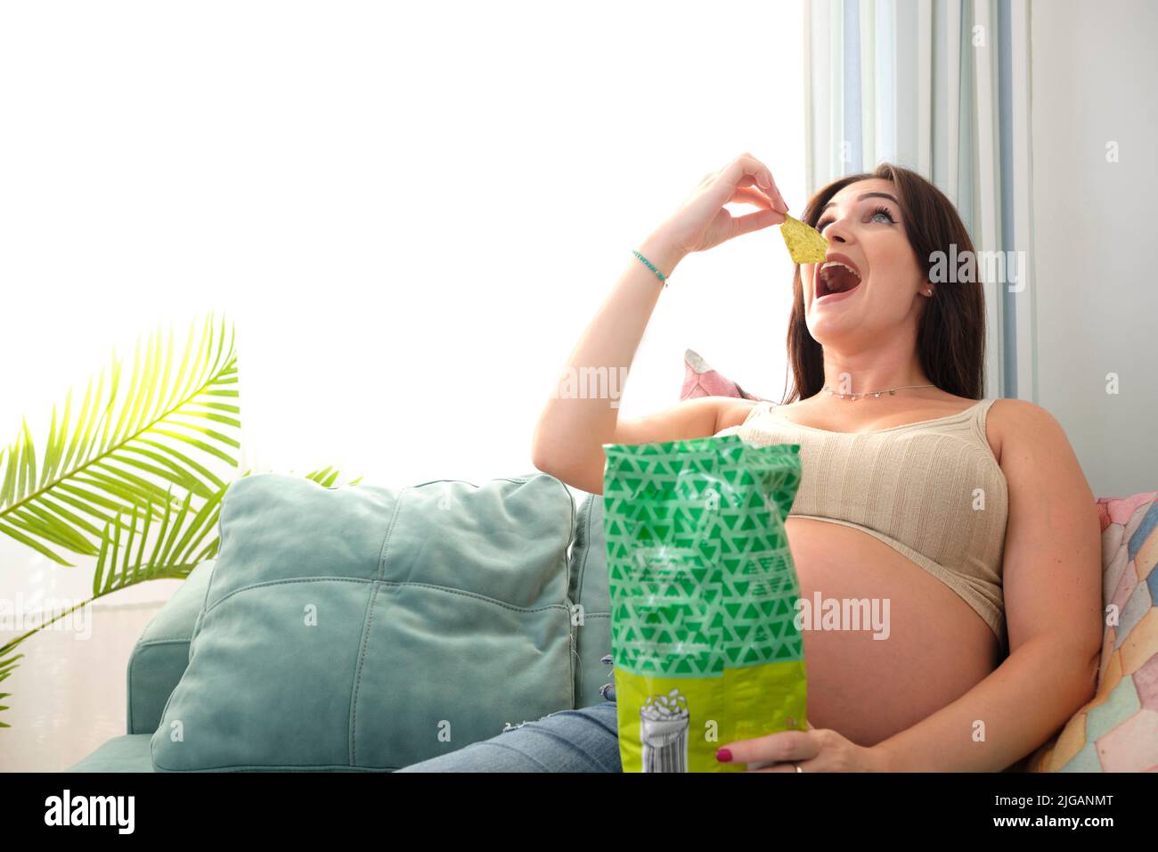 Pregnant young woman eating chips on the sofa at home. Food and
