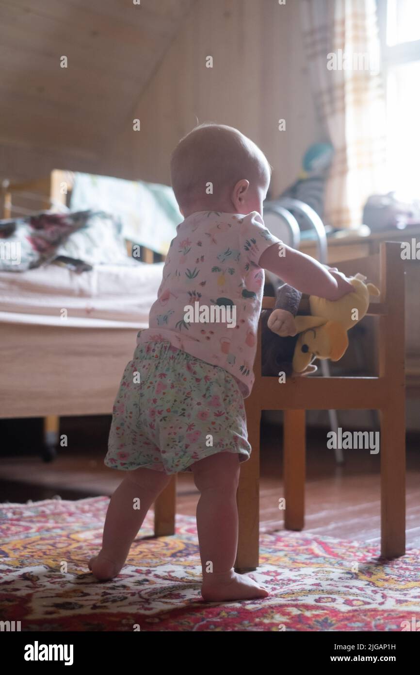 Little baby girl stands or climbs on the chair in the room. the baby is learning to walk Stock Photo