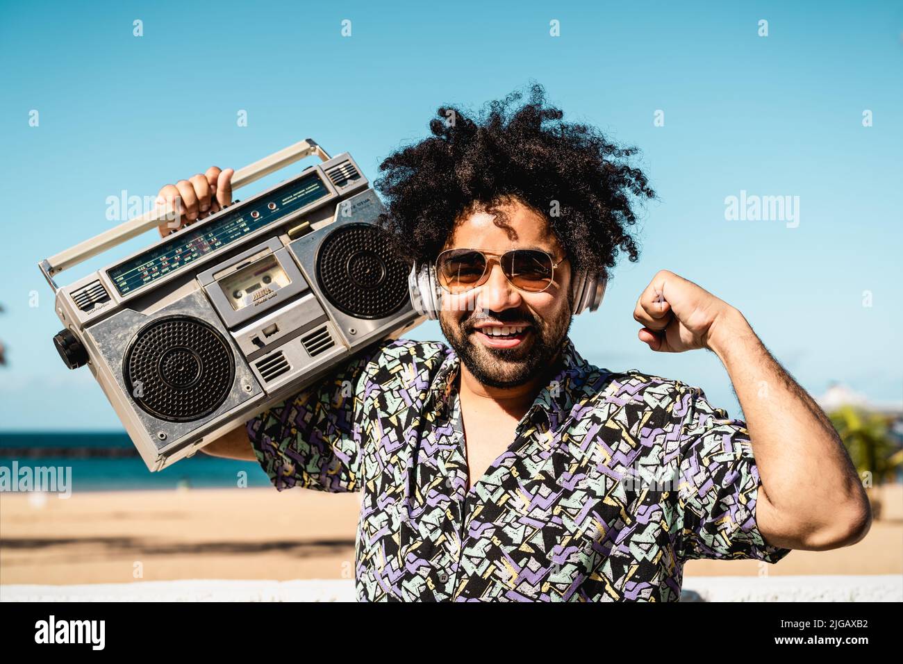 Happy Latin man having fun listening to music with wireless headphones and vintage boombox on the beach during summer time Stock Photo