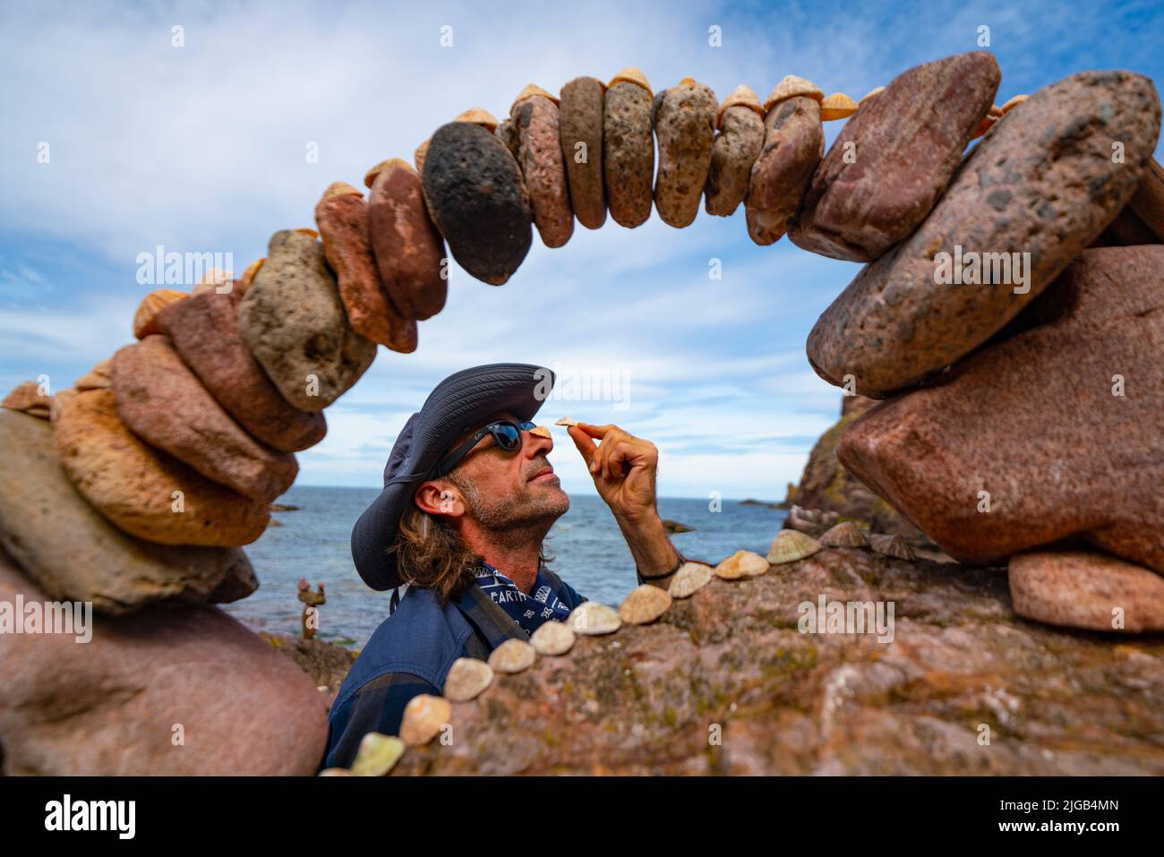 Dunbar, Scotland, UK. 9 July 2022. Day one of the 11th Stone Stacking Championships held at Eye Cave Beach in Dunbar in East Lothian. . Competitors are shown during the arch building competition. Pic; Pedro Duran stacking seashells on his nose beside his stone arch. Iain Masterton/Alamy Live News Stock Photo