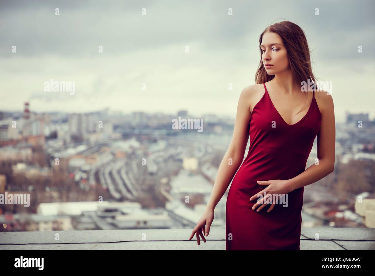 Slim girl in red dress is standing on top of skyscraper, high above the megacity, sprawling behind her till the horizon. Stock Photo