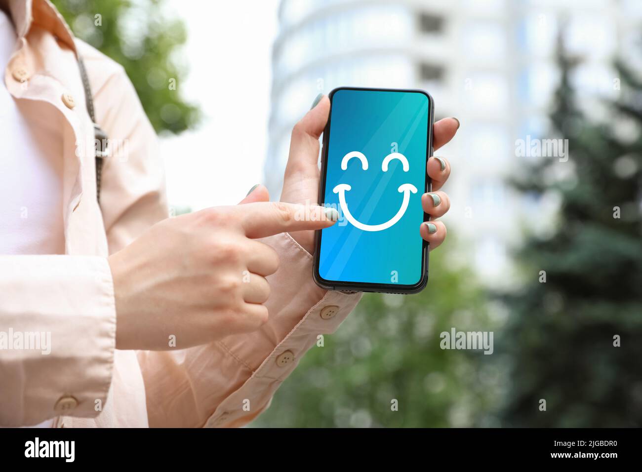 Woman holding phone with happy emoticon on screen outdoors, closeup Stock Photo