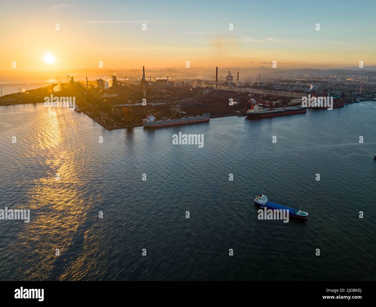 Cargo ships docked at large industrial facility with smokestacks at sunset Stock Photo
