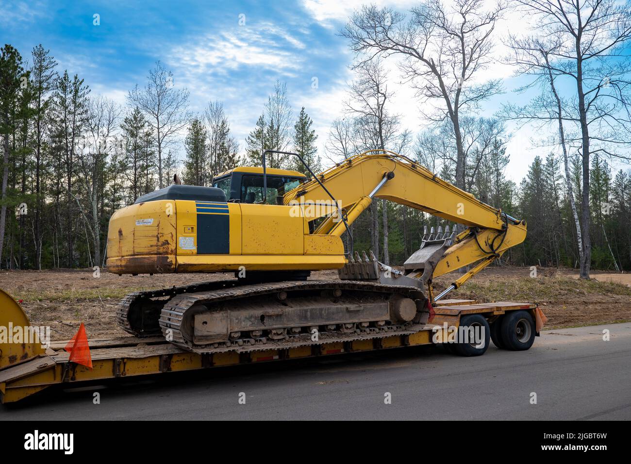 Used Yellow Skid Steer Excavator Loader on a flat bed trailer, at a construction job site, in evening light with trees in the background. Stock Photo