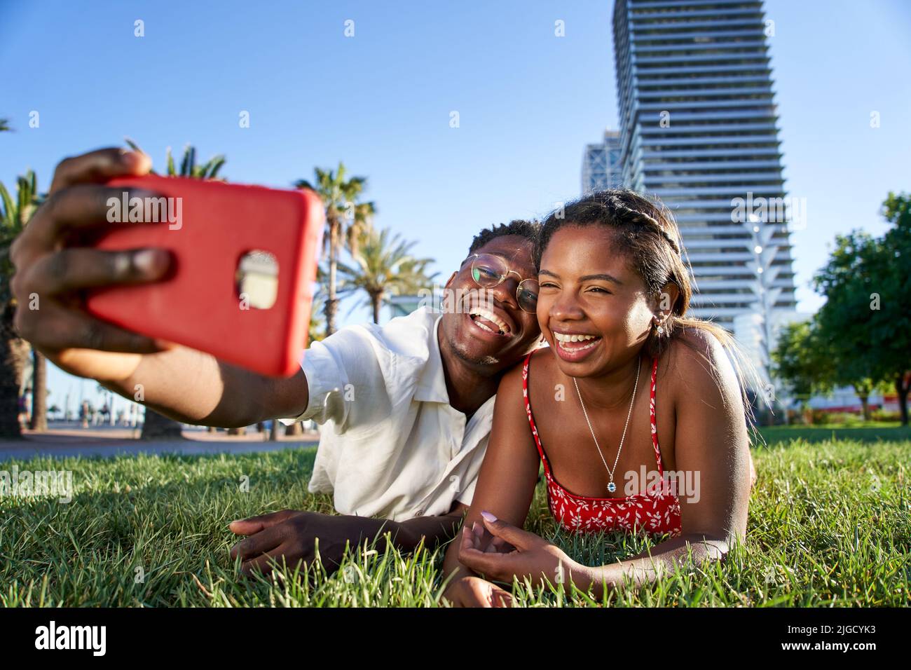 A cheerful young African American couple taking a selfie portrait with their smart phone lying on the grass in the city park. Smiling boy and girl Stock Photo