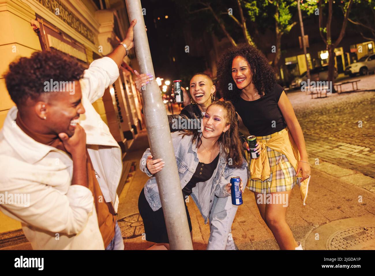 Playful friends having fun in the city at night. Group of carefree young friends laughing happily while standing around a pole. Multicultural young pe Stock Photo