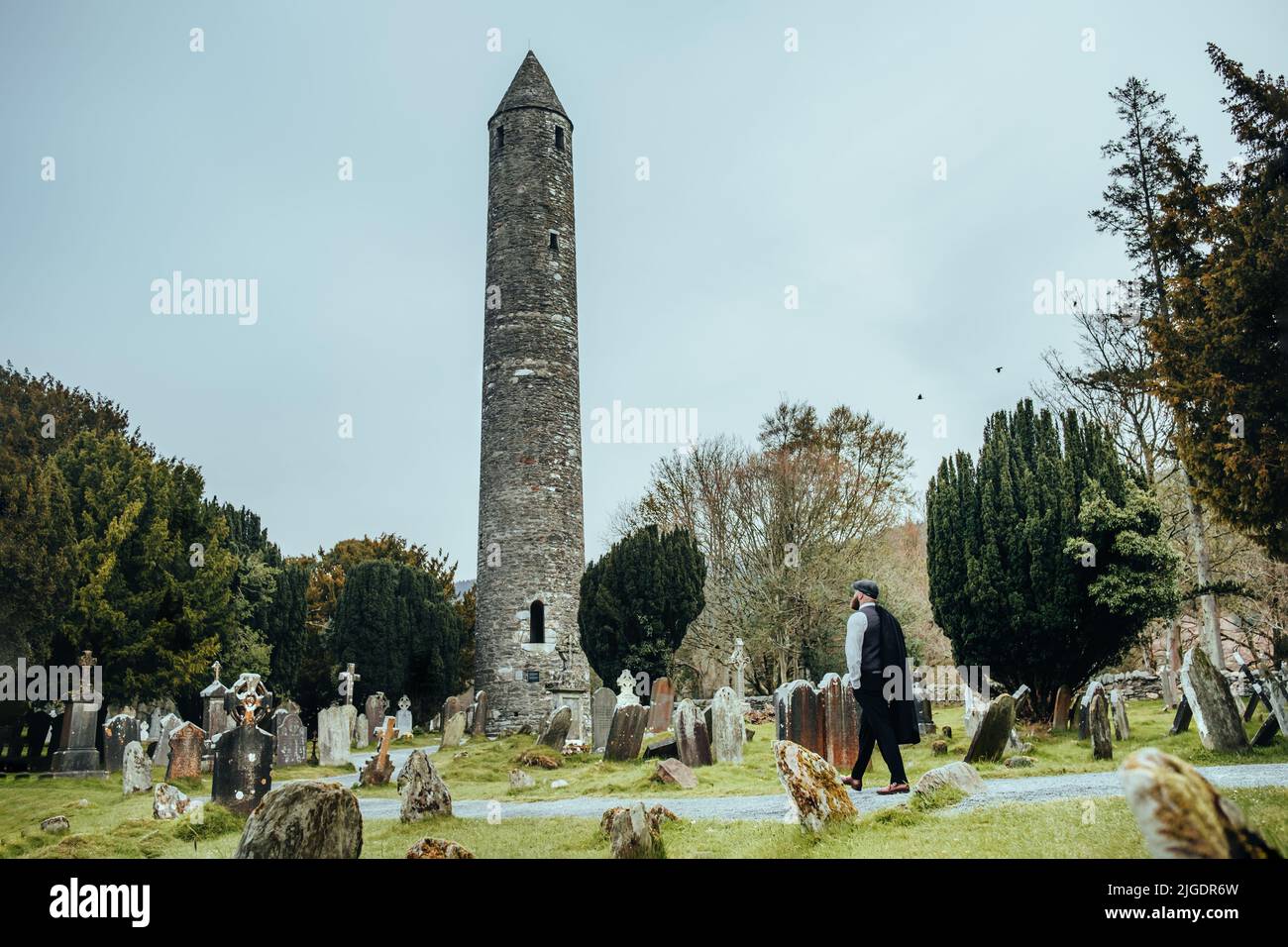 Stylish man walking towards ancient ruins & old cemetery Wicklow mountains, Ireland. 1920s theme. Fashionable brutal confident bearded man. Stock Photo