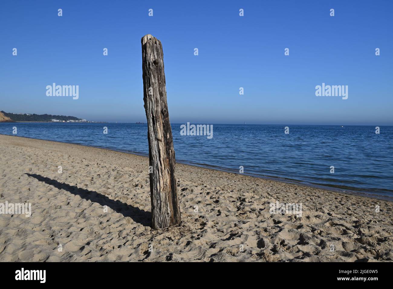 Old log, standing upright, stuck in the sand at a footprint covered beach, with calm sea water lapping the shore in the background Stock Photo