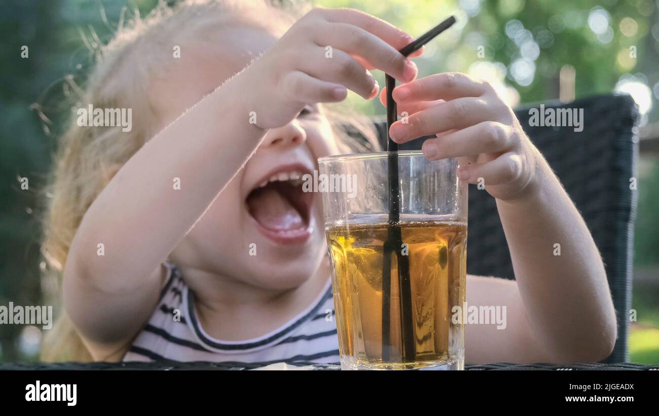 Cute little girl drinks juice through straw. Close-up portrait of blonde girl drinks juice from glass through cocktail straw sitting in street cafe on Stock Photo