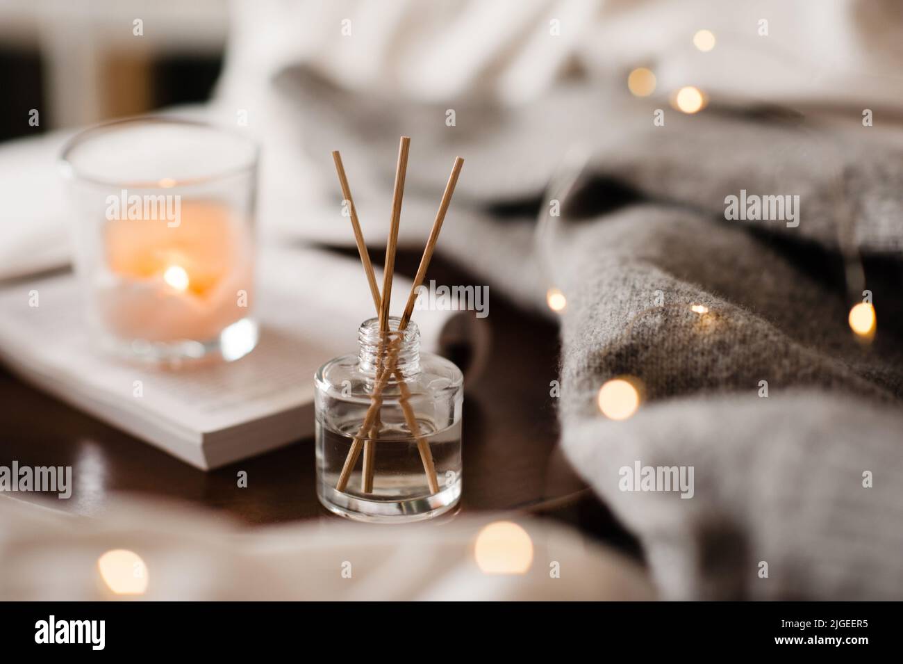 Home liquid fragrance on open paper book and burning scented candle on wooden tray and knit cloth sweater in bed close up. Cozy atmosphere. Stock Photo