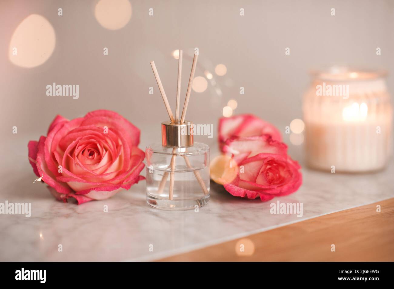 Liquid scented home fragrance in glass bottle and fresh rose flowers over burning candle and glow lights in room. Cozy atmosphere. Stock Photo