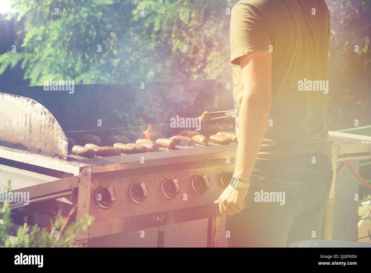 Guy cooking sausages on barbecue grill, open fire on a summer day in the backyard of a private house. Stock Photo