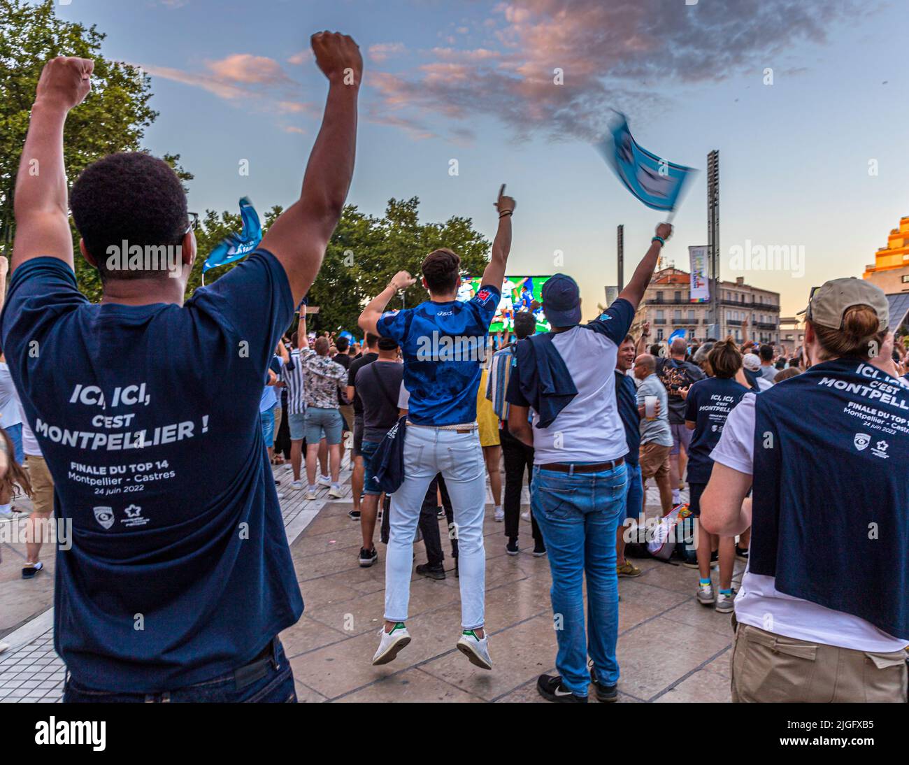 Fans raise their fists in celebration during the public viewing of the Top 14 rugby final on the Place de la Comédie in Montpellier, France. A shirt in the foreground reads "Ici, ici, c'est Montpellier!" and names the final, Montpellier against Castres, on 24 June 2022. Montpellier Hérault Rugby beat Castres Olympique 29–10 in the match played at the Stade de France in Paris, winning the club's first French championship title. Stock Photo