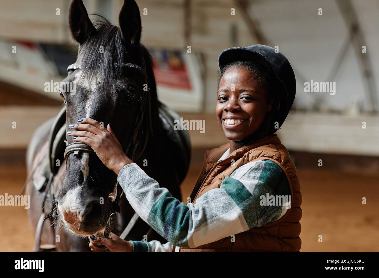 Waist up portrait of smiling black woman posing with horse in indoor riding arena and looking at camera, copy space Stock Photo