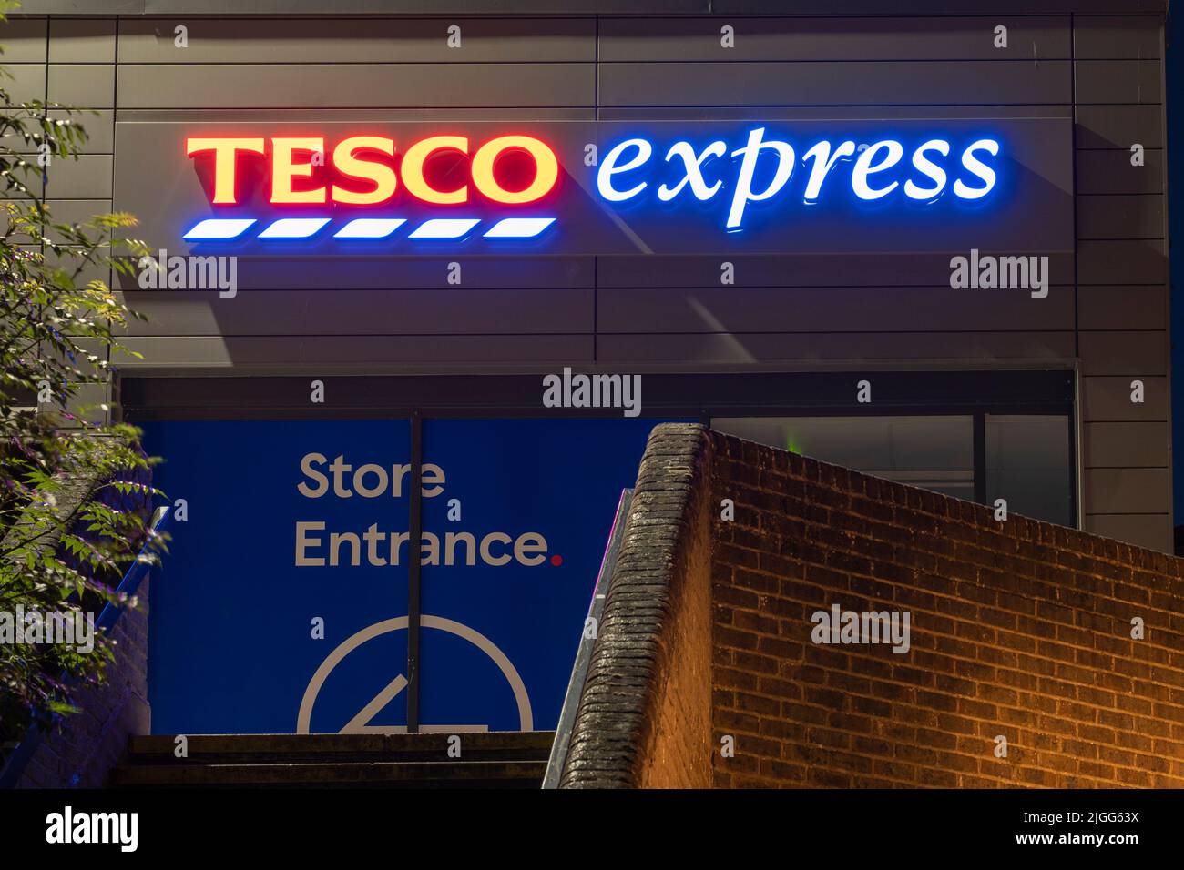 Tesco Express - a small grocery supermarket - logo and sign illuminated at night in The Malls shopping centre in Basingstoke town centre. England, UK Stock Photo