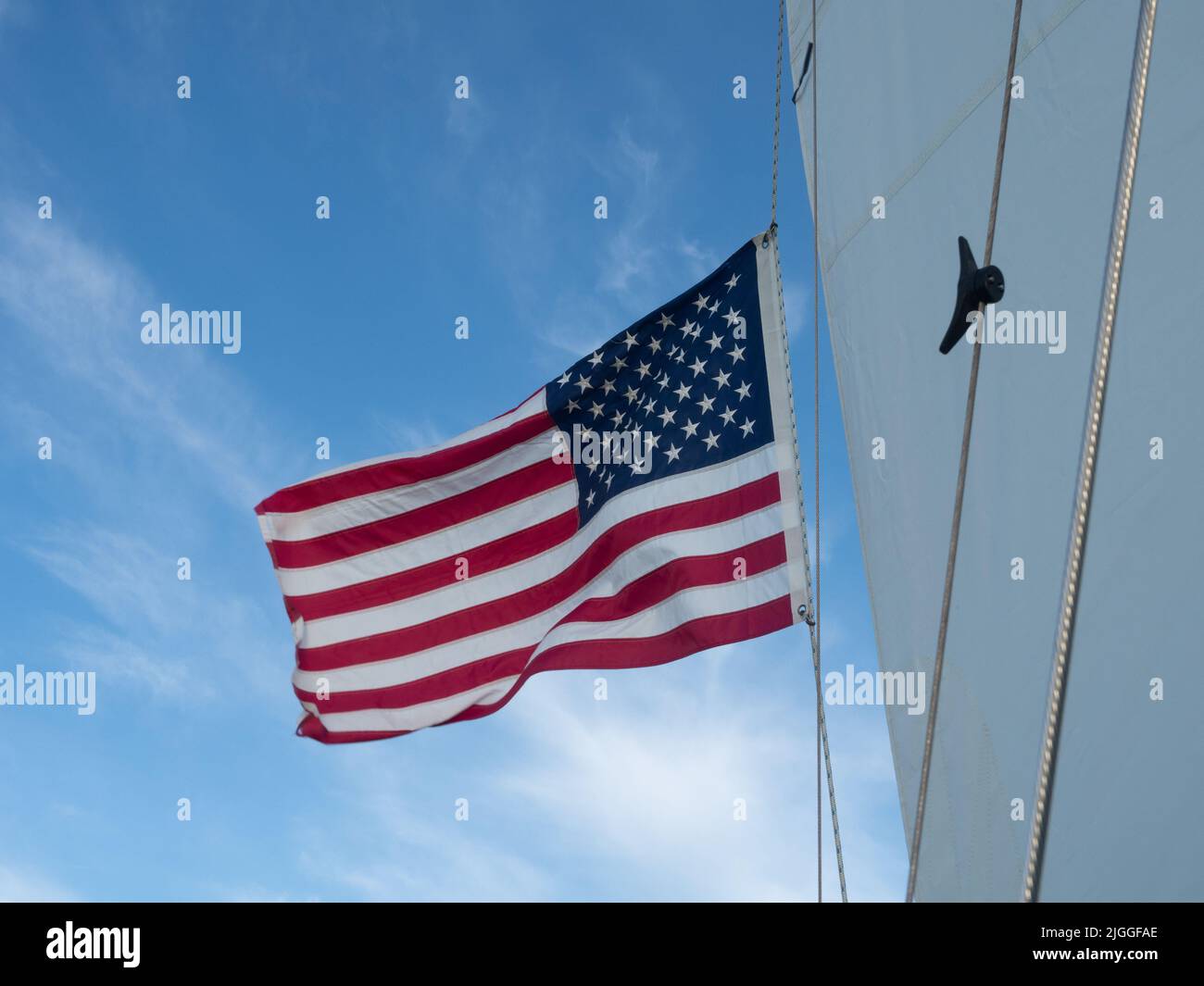 An unfurled American flag flying from a mast or sailboat rig with blue sky and thin clouds in background. Stock Photo