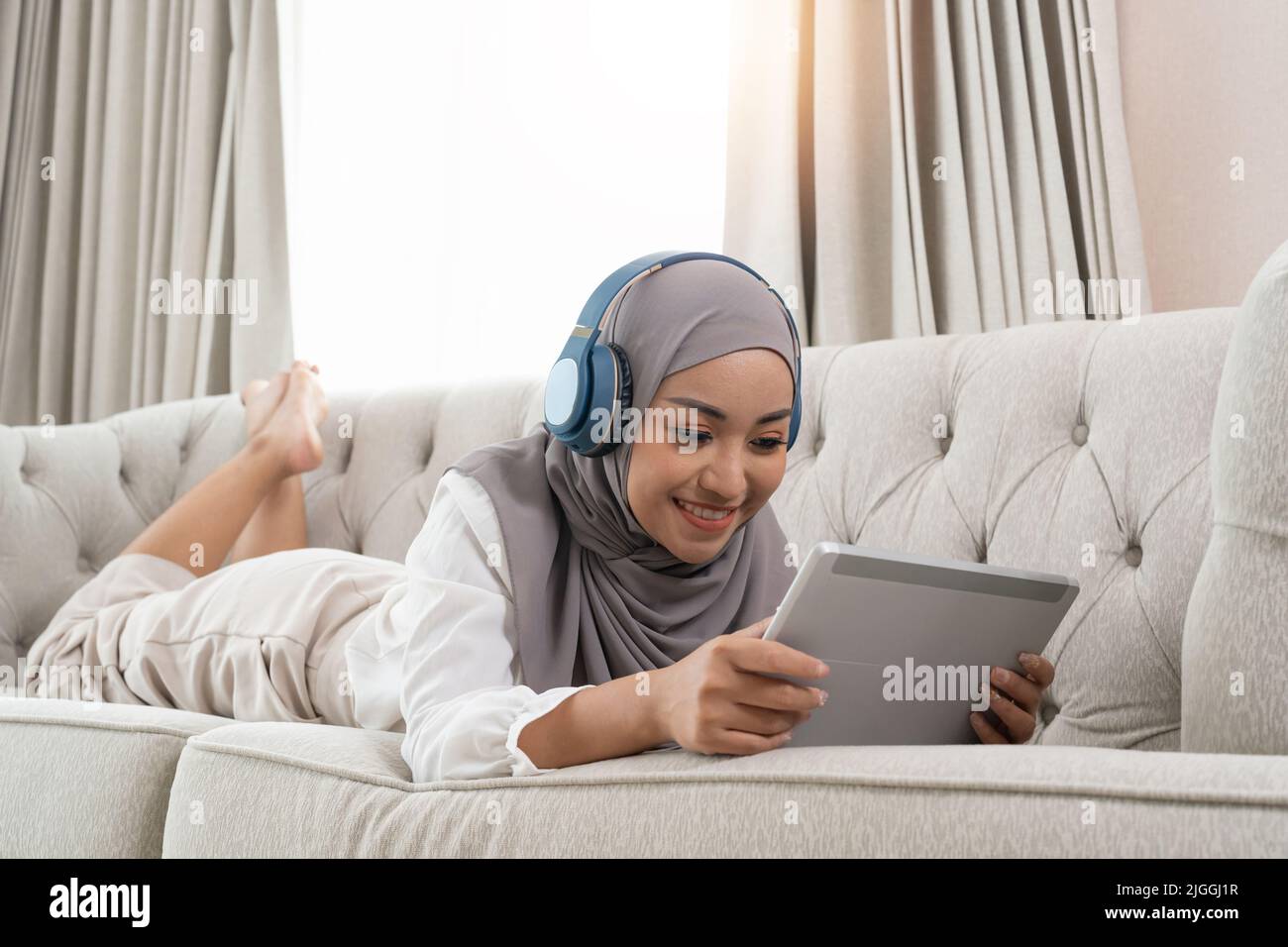 full length view of young muslim woman laying on couch in living room and watching movie on digital tablet Stock Photo