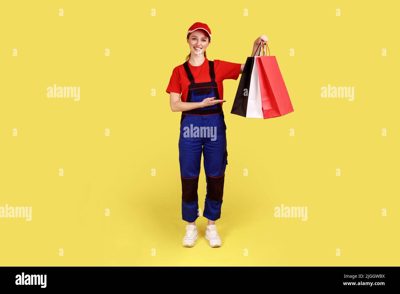 Beautiful optimistic courier woman standing with shopping bags in hands, showing presenting parcels, expressing happiness, wearing overalls and cap. Indoor studio shot isolated on yellow background. Stock Photo