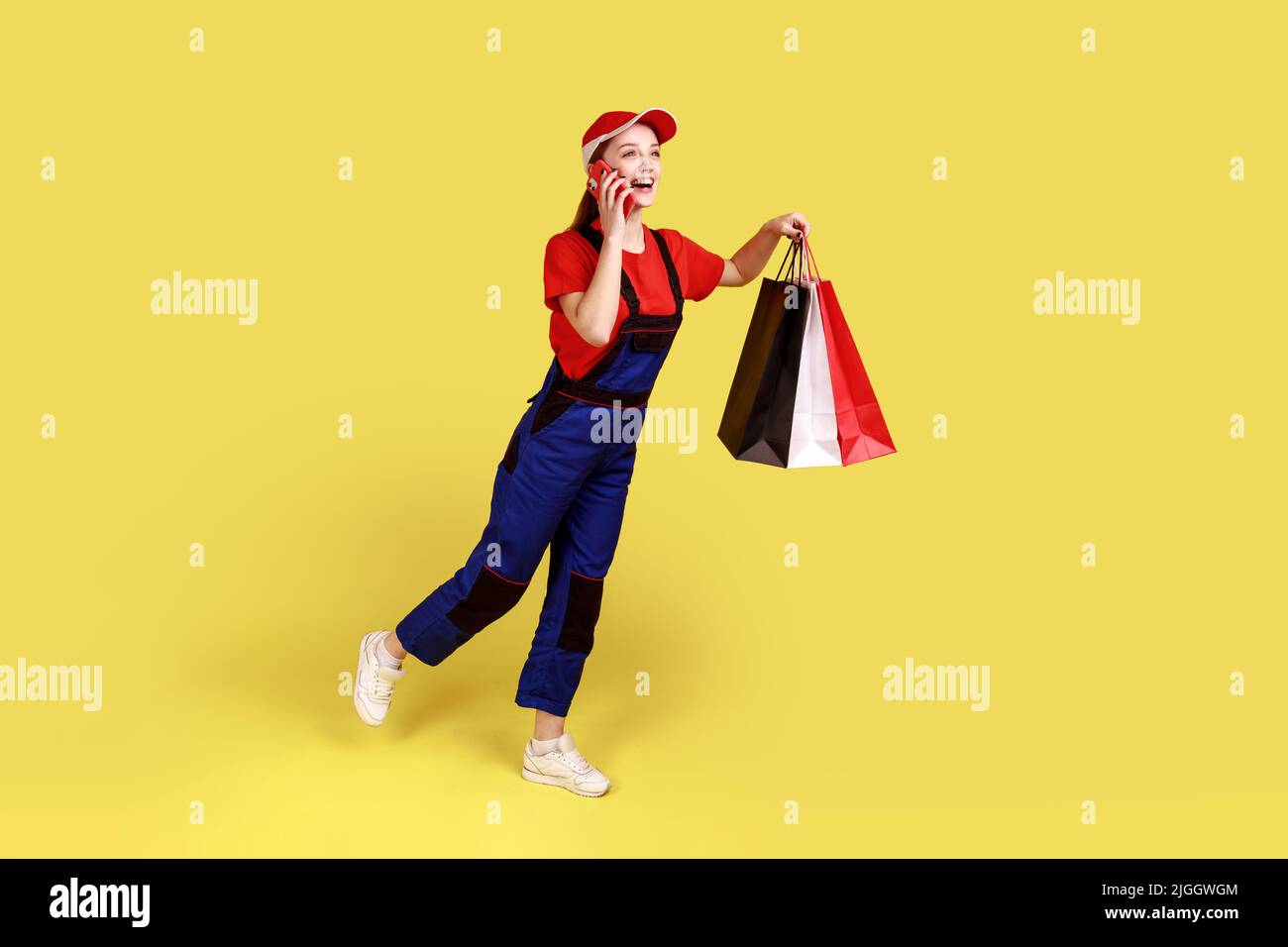 Full length portrait of excited satisfied courier woman being hurry to deliver shopping bags to client, wearing overalls and red cap. Indoor studio shot isolated on yellow background. Stock Photo