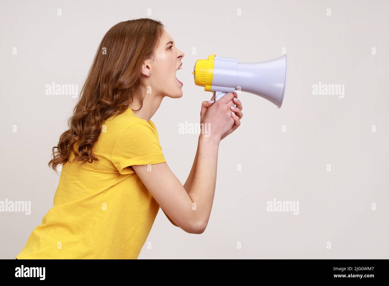 Profile portrait nervous brown haired female of young age in yellow t-shirt loudly speaking screaming holding megaphone, announcing important message. Indoor studio shot isolated on gray background. Stock Photo