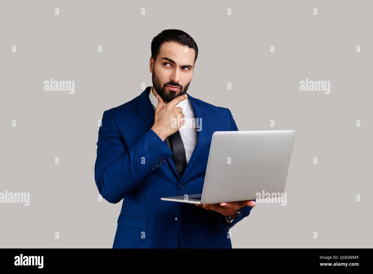 Thoughtful bearded man holding laptop and thinking over startup strategy with serious doubtful expression, wearing official style suit. Indoor studio shot isolated on gray background. Stock Photo
