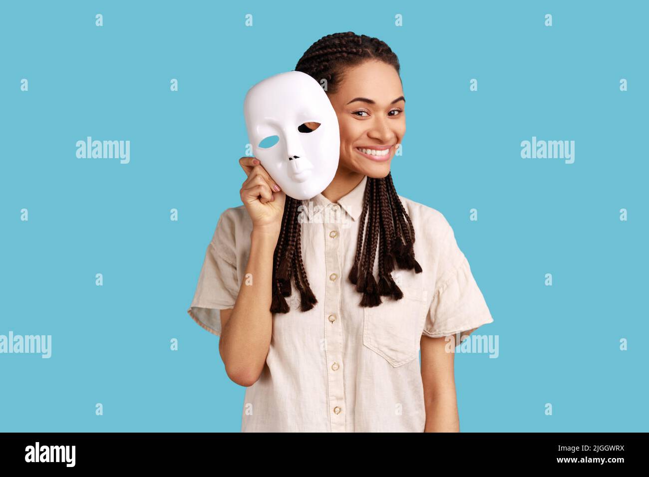 Woman with dreadlocks removing white mask from face showing his smiling expression, good mood, pretending to be another person, wearing white shirt. Indoor studio shot isolated on blue background. Stock Photo