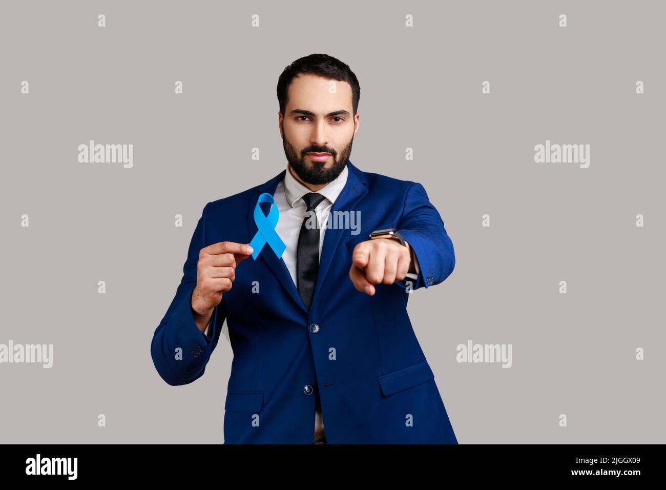 Serious confident bearded man holding blue ribbon, symbol of Colorectal cancer awareness, pointing finger ant camera, wearing official style suit. Indoor studio shot isolated on gray background. Stock Photo