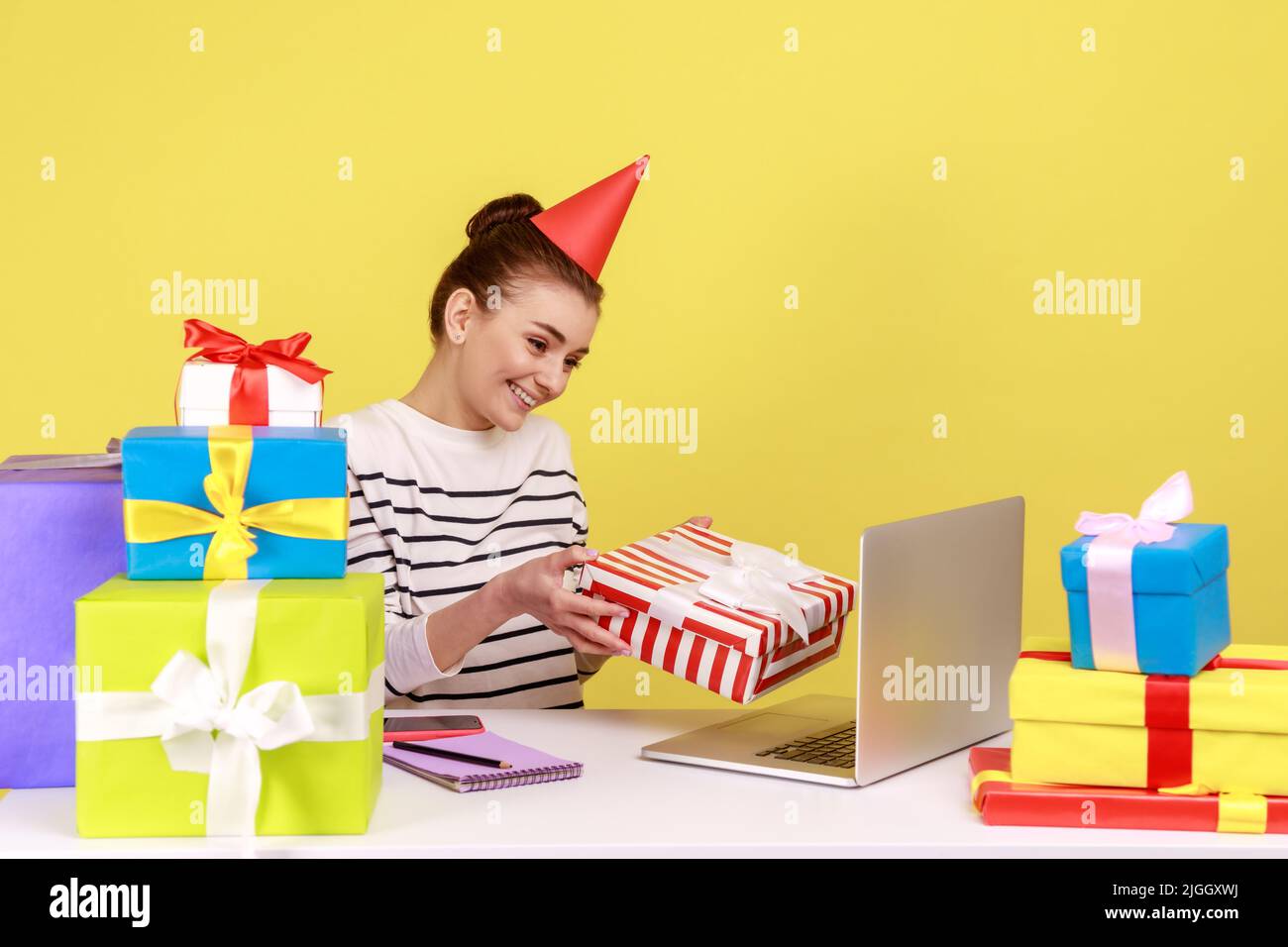 Cheerful woman holding gift box and looking at laptop screen, giving birthday present while having online communication, distant celebration. Indoor studio studio shot isolated on yellow background. Stock Photo