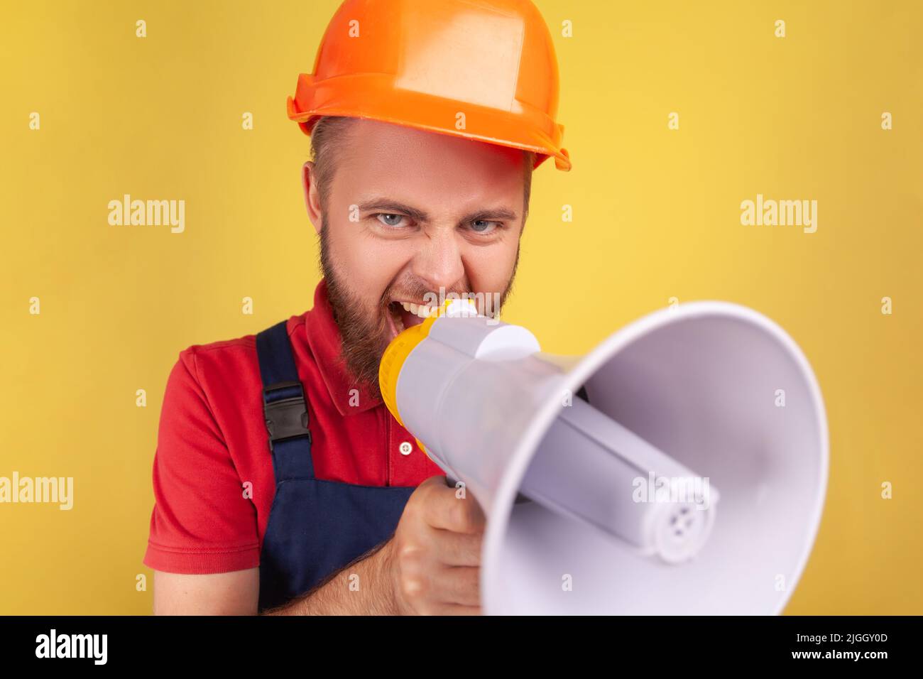 Angry aggressive bearded worker wearing protective helmet and blue overalls holding megaphone and screaming, point of view photo. Indoor studio shot isolated on yellow background. Stock Photo